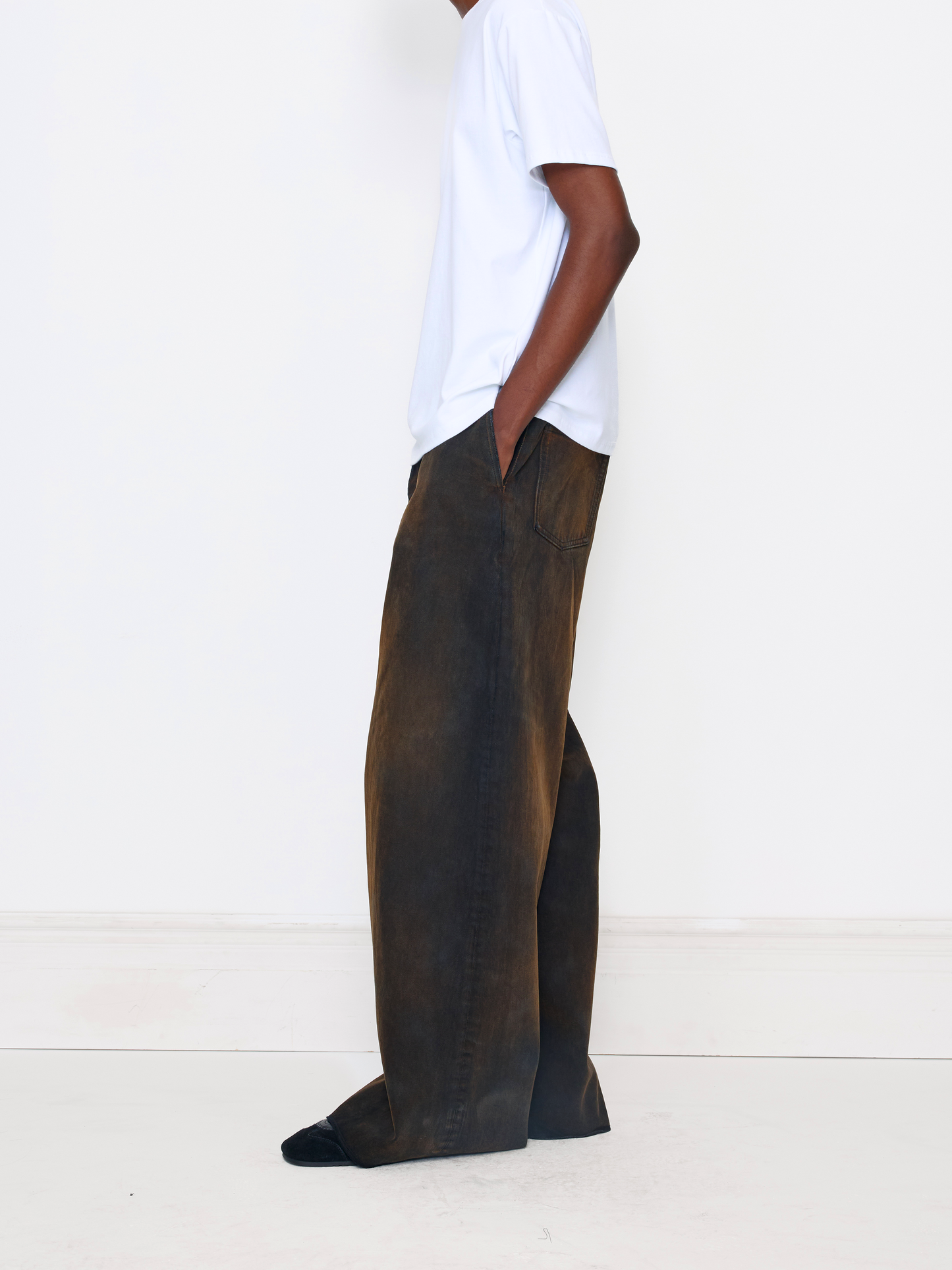 A model shown from the side wearing dark brown wide-leg trousers, a white t-shirt, and black slip-on shoes against a plain white studio background.