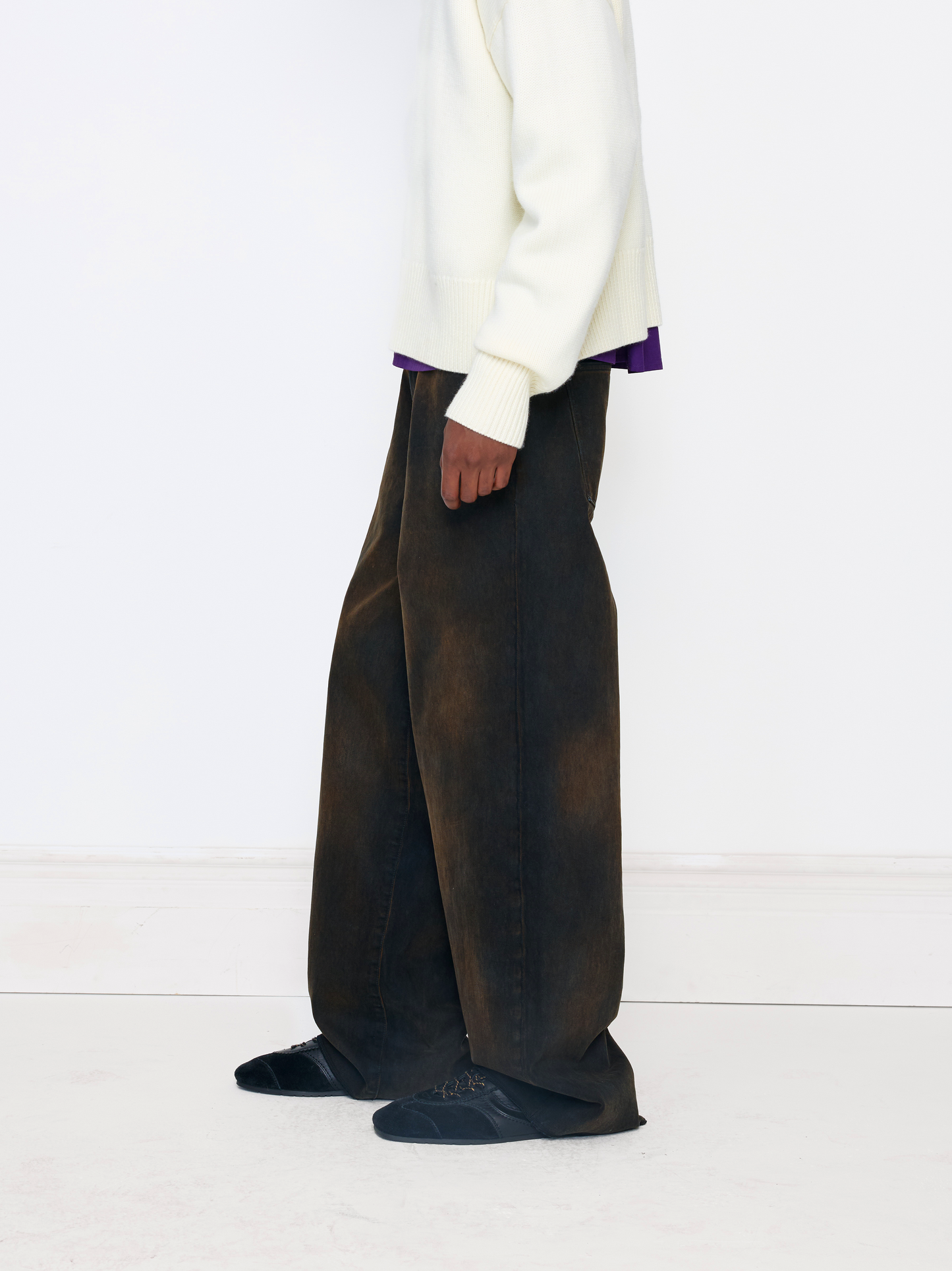 A person shown from the waist down wearing very wide-leg dark brown pants with a subtle vertical gradient, paired with a cream sweater and black shoes, against a clean white studio background.