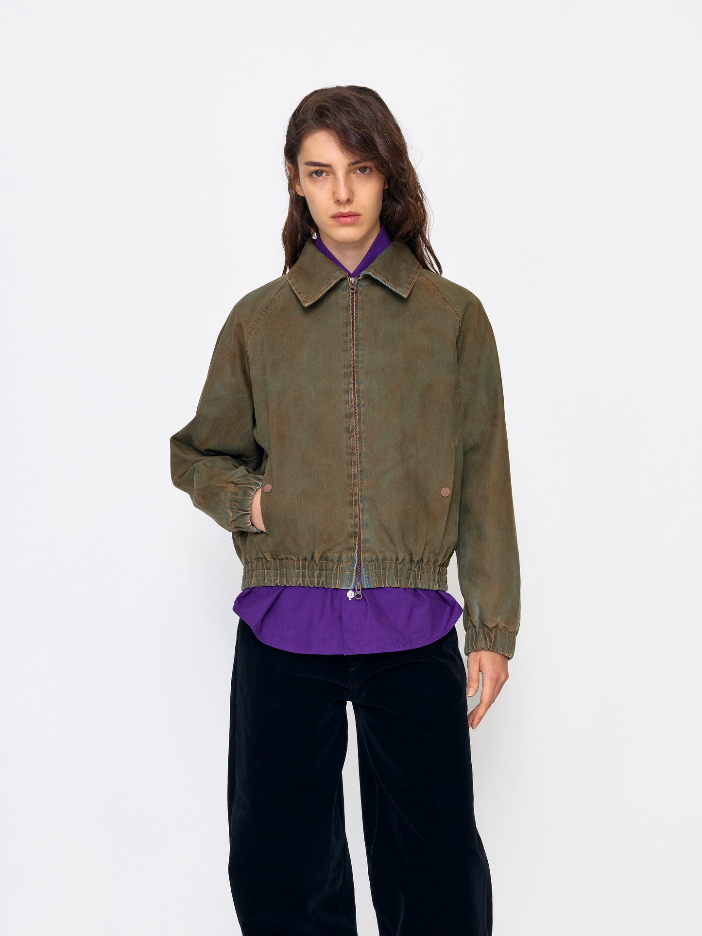 A model stands against a plain white studio background wearing a cropped olive-brown suede jacket over a purple collared shirt and black pants.