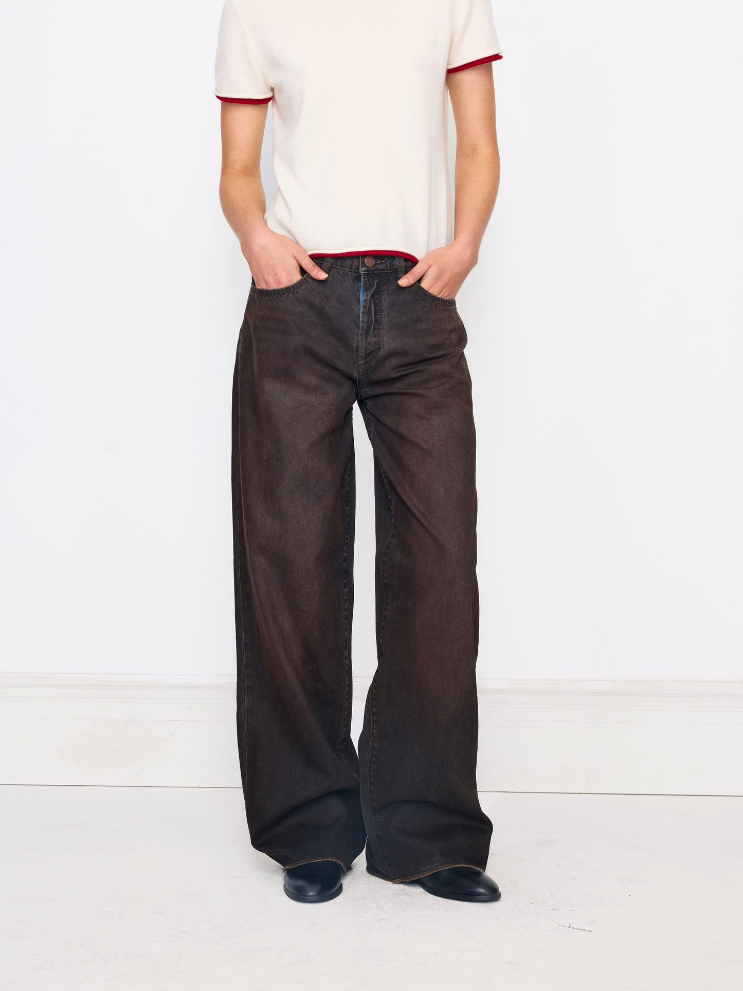 A model wearing dark brown wide-leg denim pants, paired with a cream t-shirt featuring red trim and black shoes, standing against a white studio background.