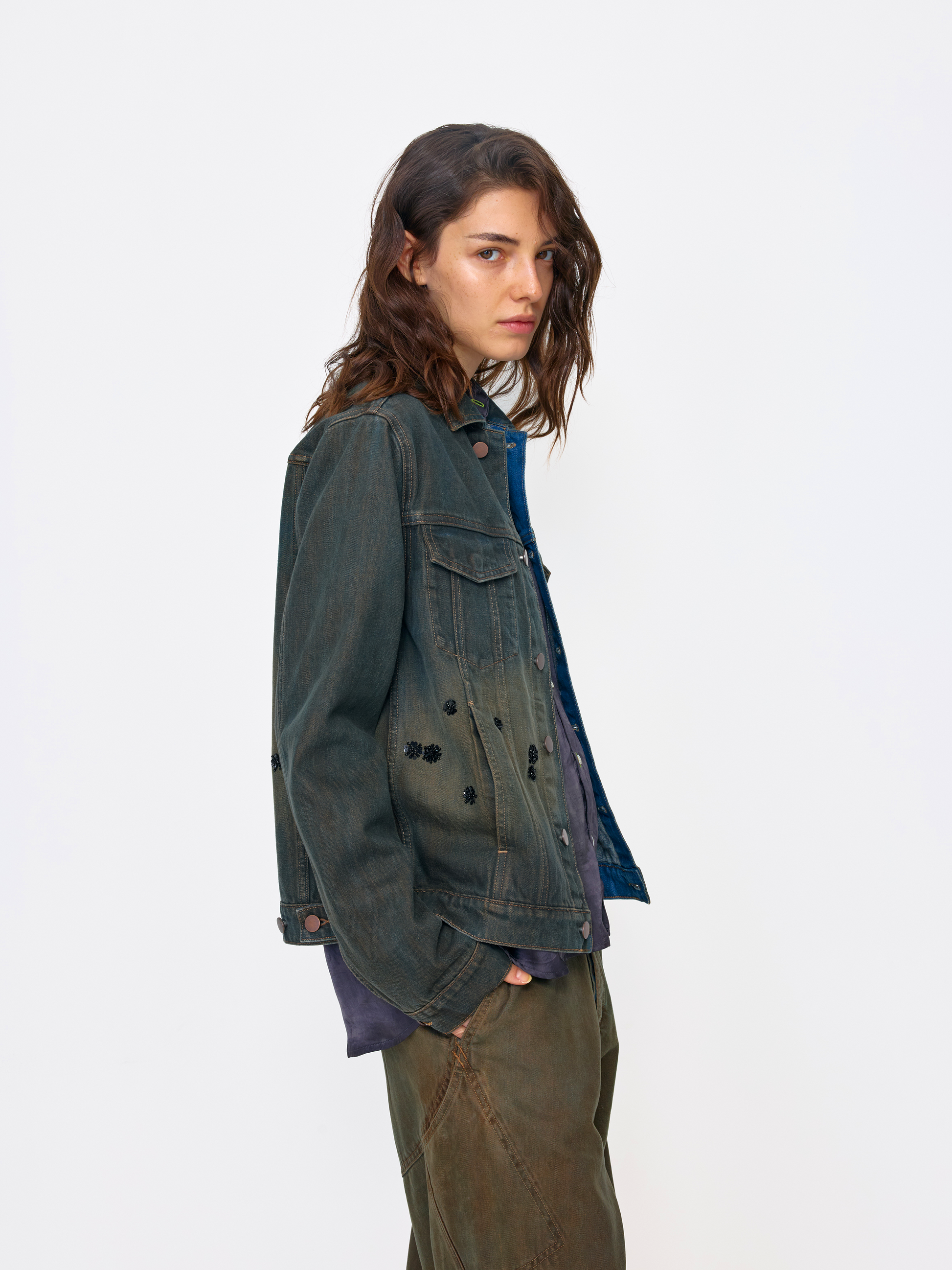 A model wearing a dark washed denim jacket over a blue shirt and olive green pants, standing against a plain white studio background.