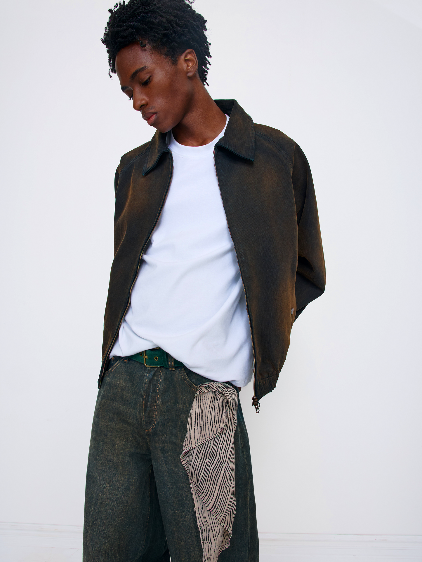 A model wearing a dark brown suede jacket over a white t-shirt, paired with dark green wide-leg jeans featuring a textured side panel and a green belt, set against a plain white studio background.
