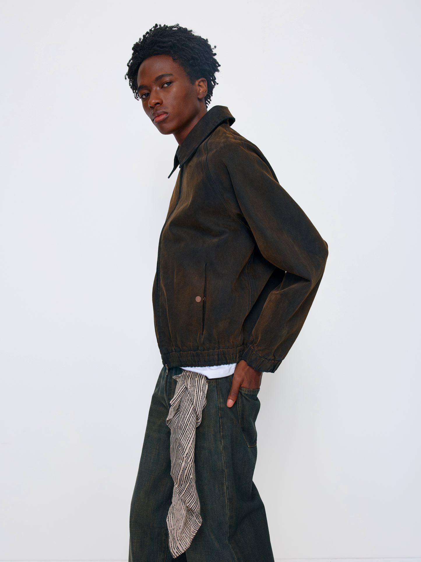 A model wearing a dark brown waxed cotton jacket, styled with dark green wide-leg jeans featuring a patterned tie detail and a white undershirt, set against a plain white studio background.