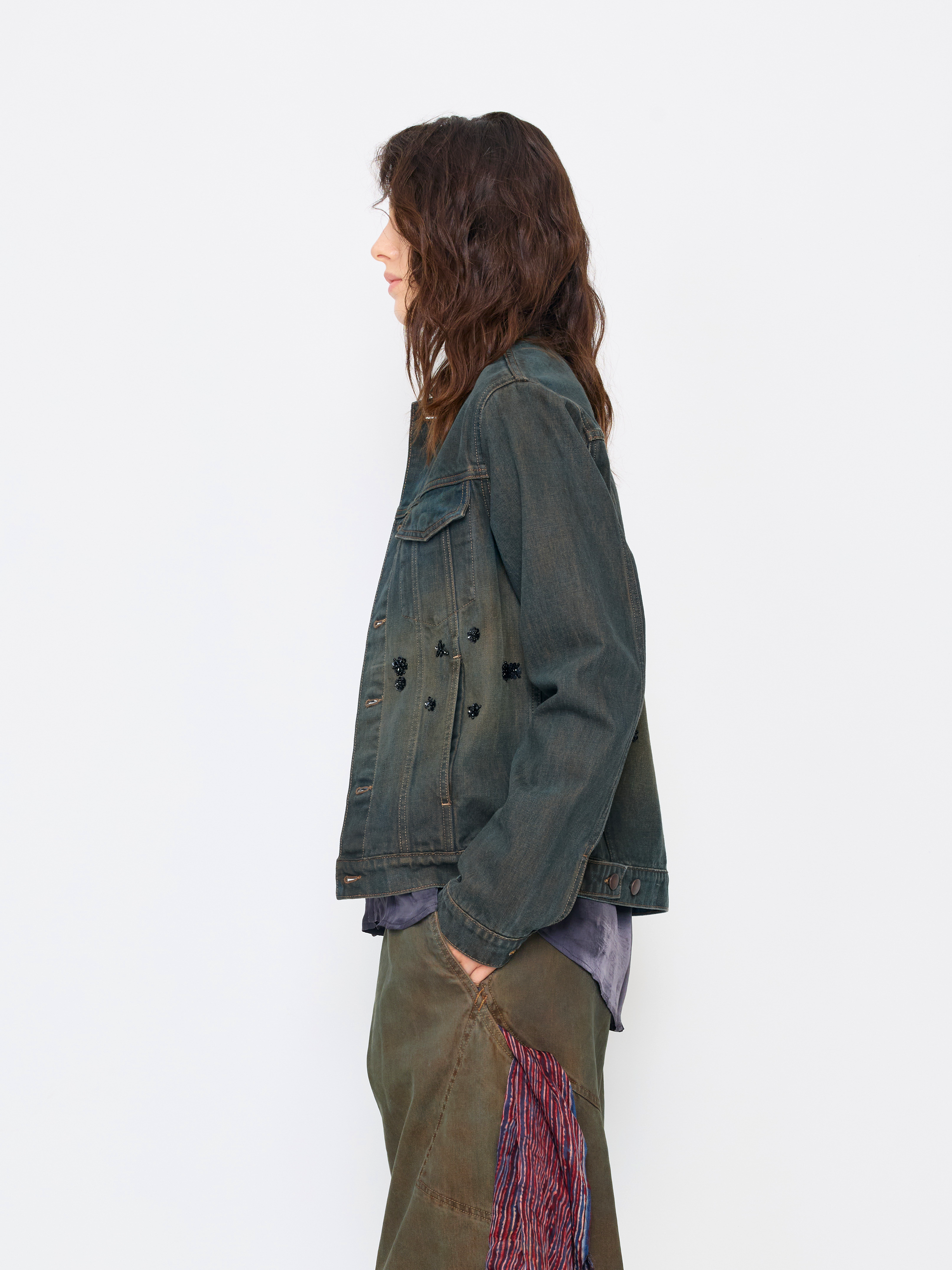 A woman shown in profile wearing a dark washed denim jacket over a layered shirt and olive green pants with a patterned side panel, photographed against a plain light background.