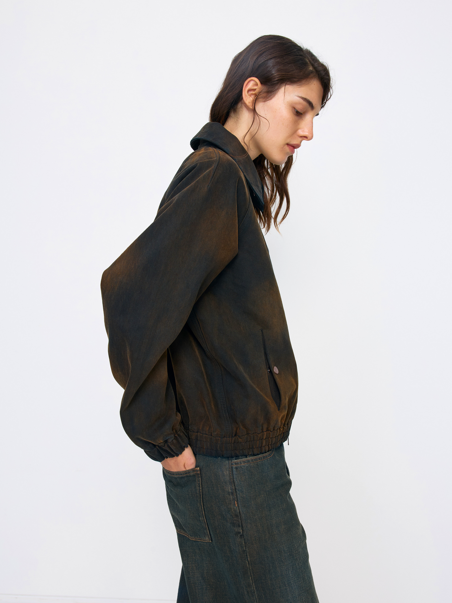 A woman shown in side profile wearing a dark brown washed jacket and dark blue jeans against a plain white studio background.