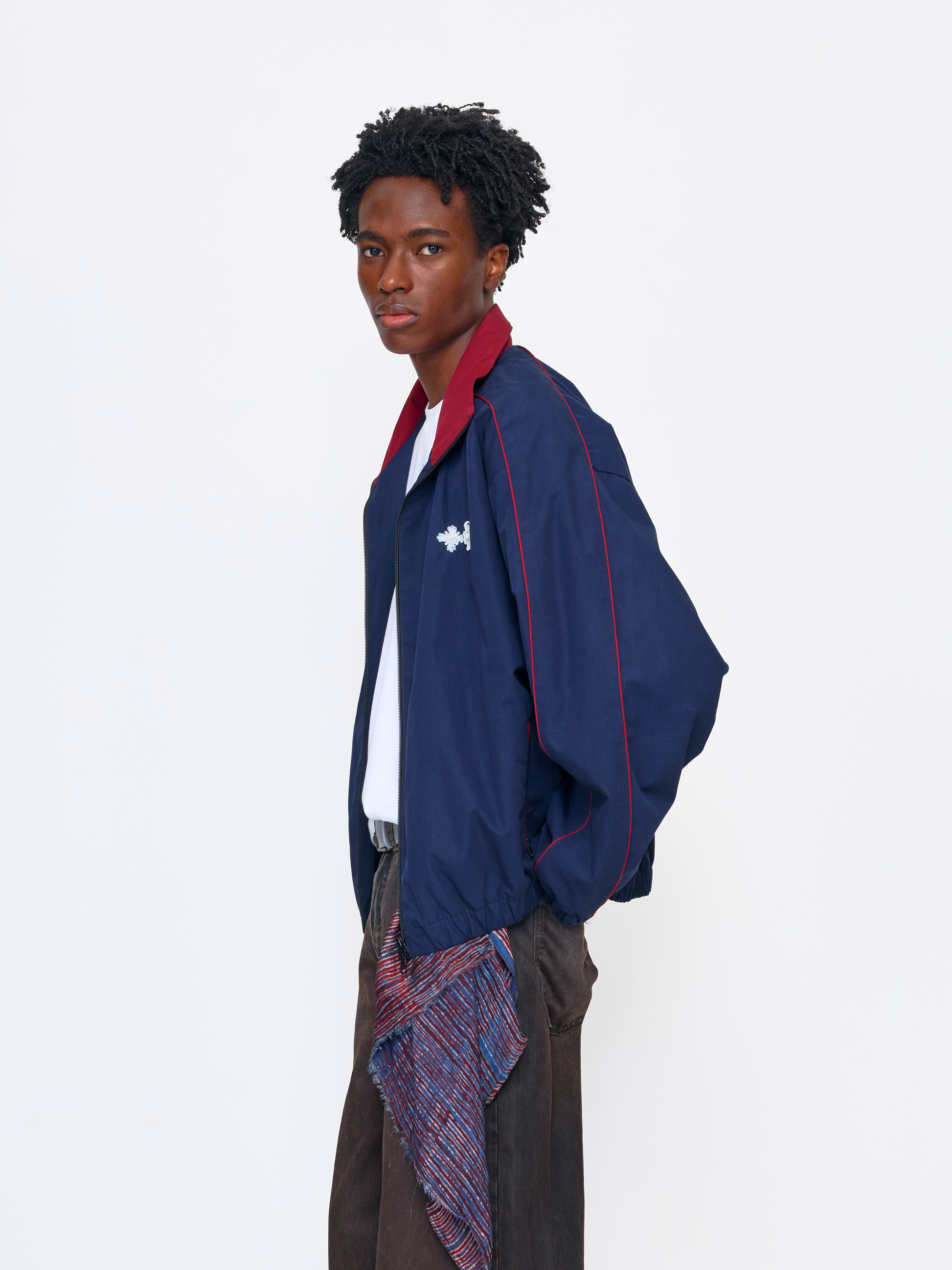 A model wearing a navy blue athletic windbreaker jacket with red trim and a white Under Armour logo, styled with a white shirt, brown trousers, and a patterned scarf, against a plain white studio background.