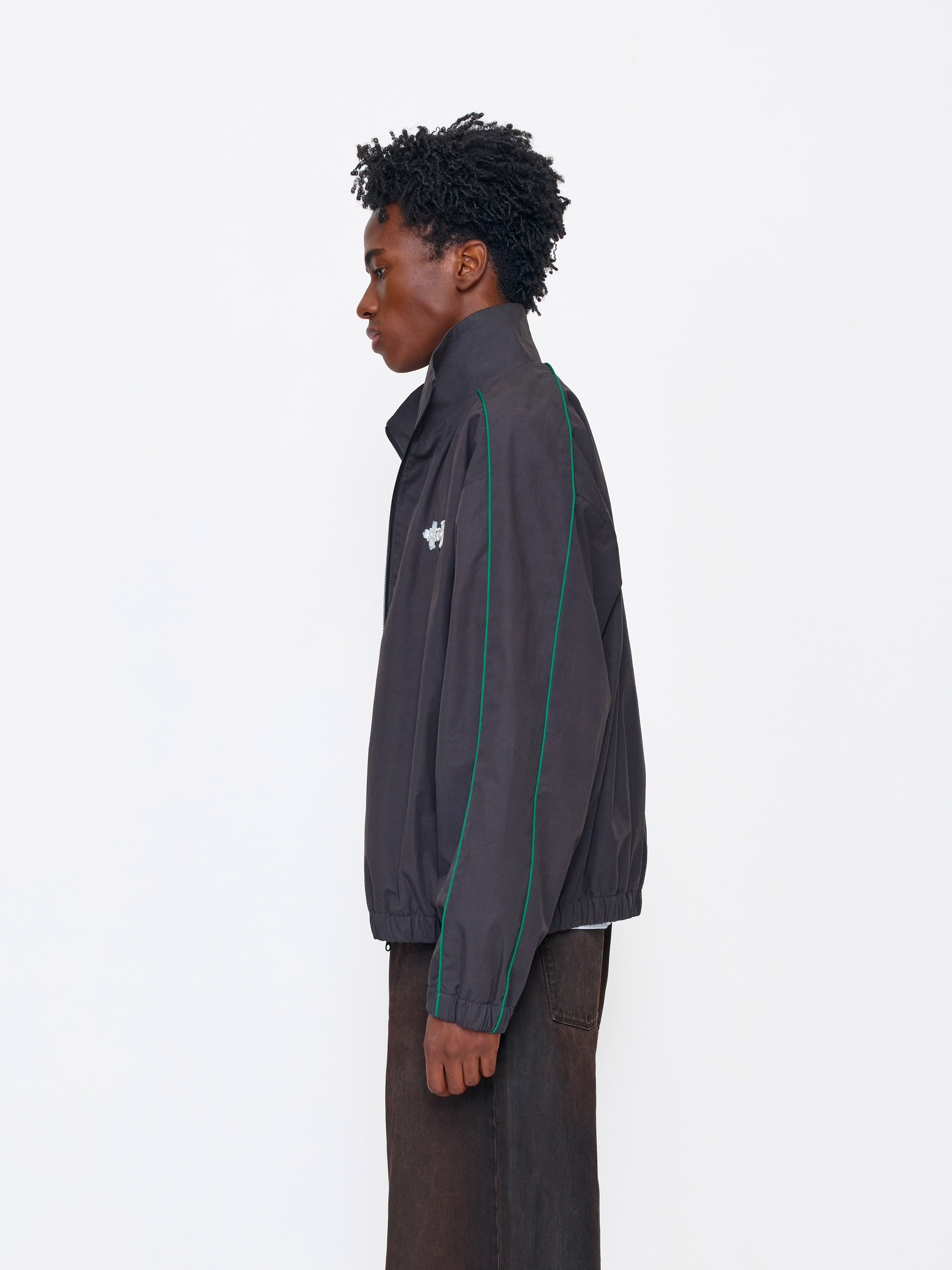 A model shown in profile wearing a dark gray zip-up windbreaker jacket with green piping along the shoulders and sleeves, paired with dark brown trousers, against a plain white studio background.