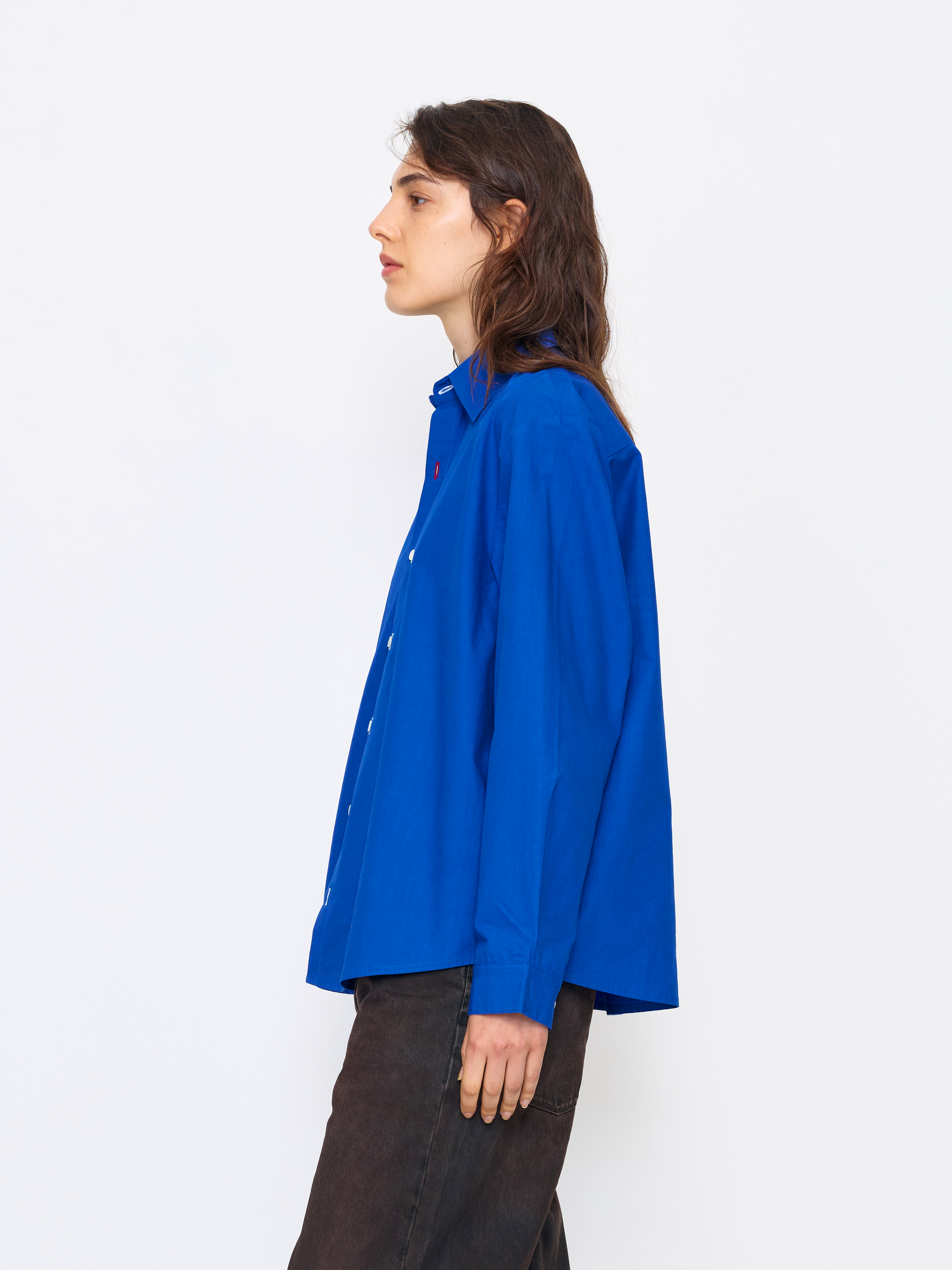 A model shown in profile wearing a vibrant royal blue long-sleeve button-up shirt and dark brown trousers, standing against a plain white studio background.