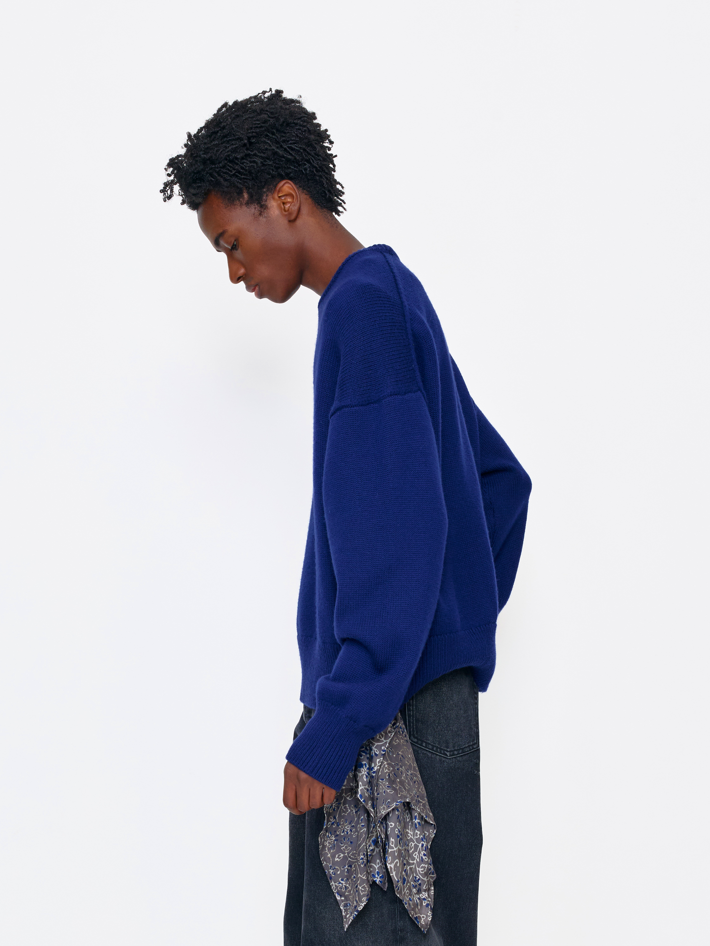 A model wearing a vibrant royal blue knit sweater, styled with dark jeans and a patterned scarf, photographed from the side against a clean white studio background.