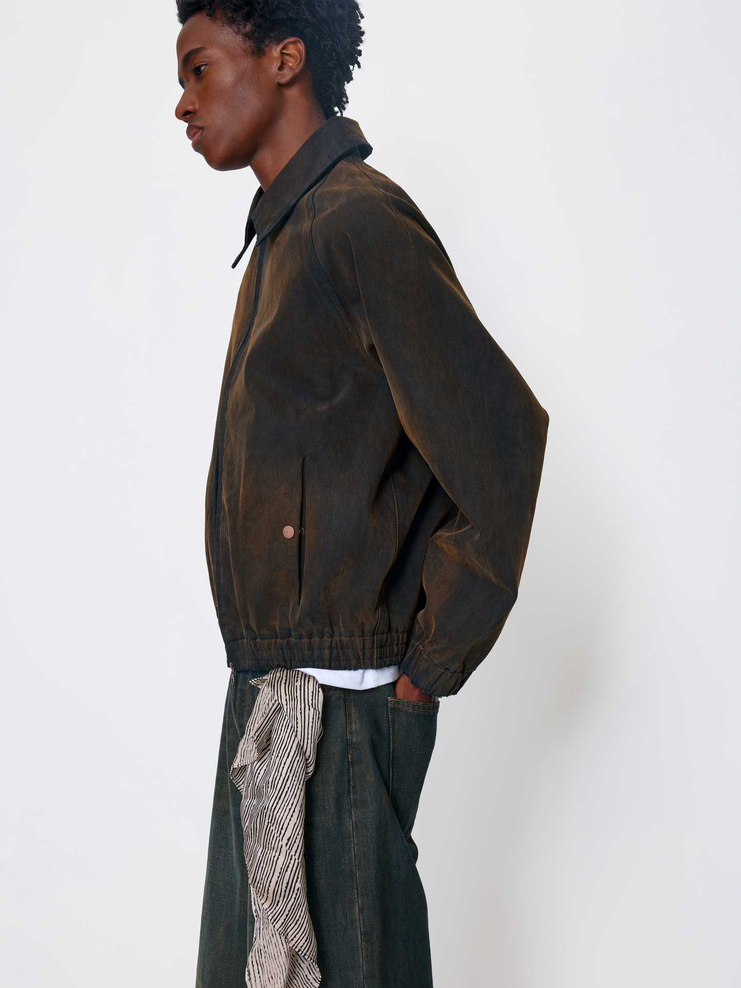 A model shown in profile wearing a dark brown corduroy jacket, dark green trousers, and a patterned shirt tucked into the pants, against a plain white studio background.