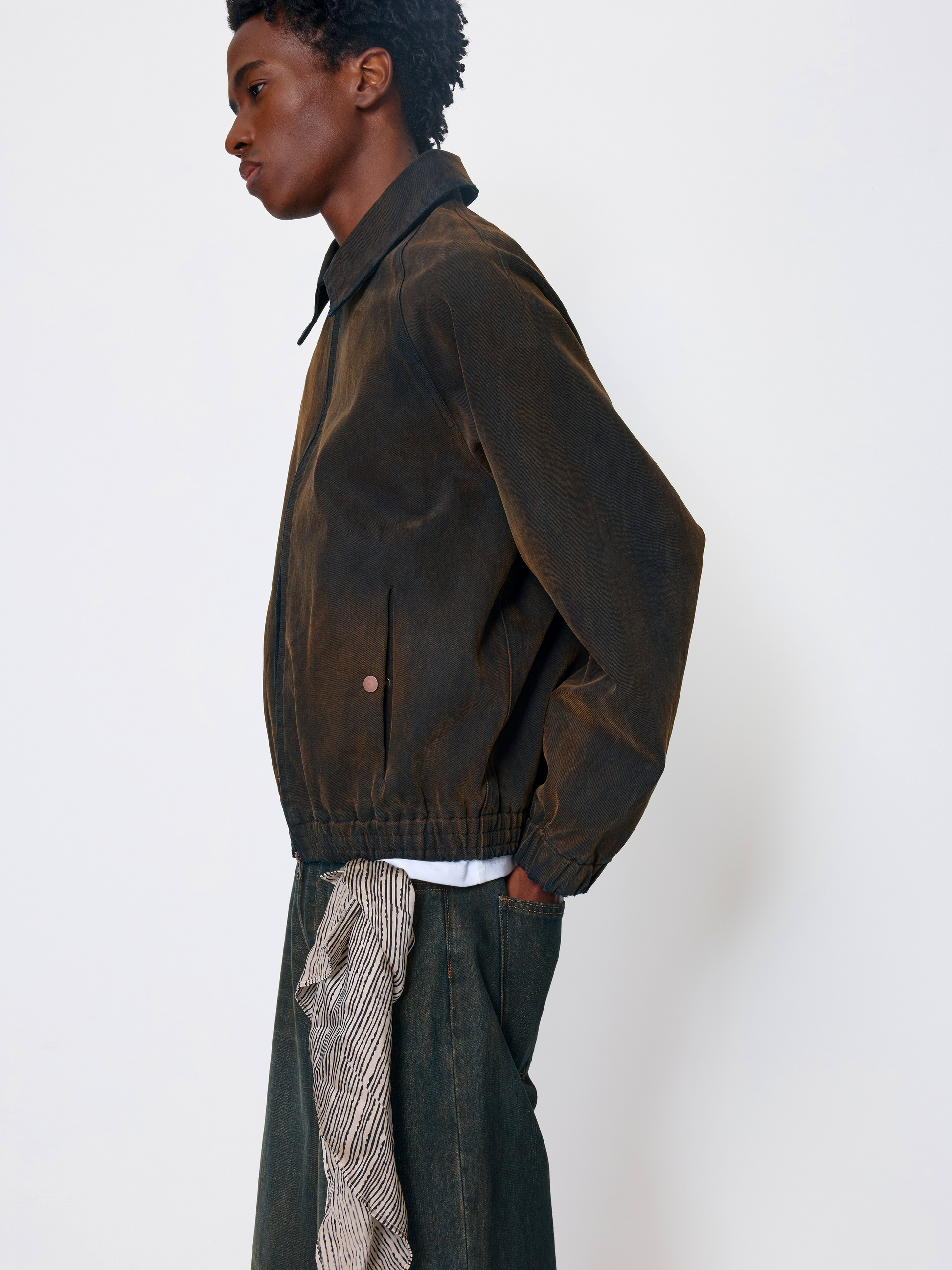A model shown in profile wearing a dark brown corduroy jacket, dark green trousers, and a patterned shirt tucked into the pants, against a plain white studio background.