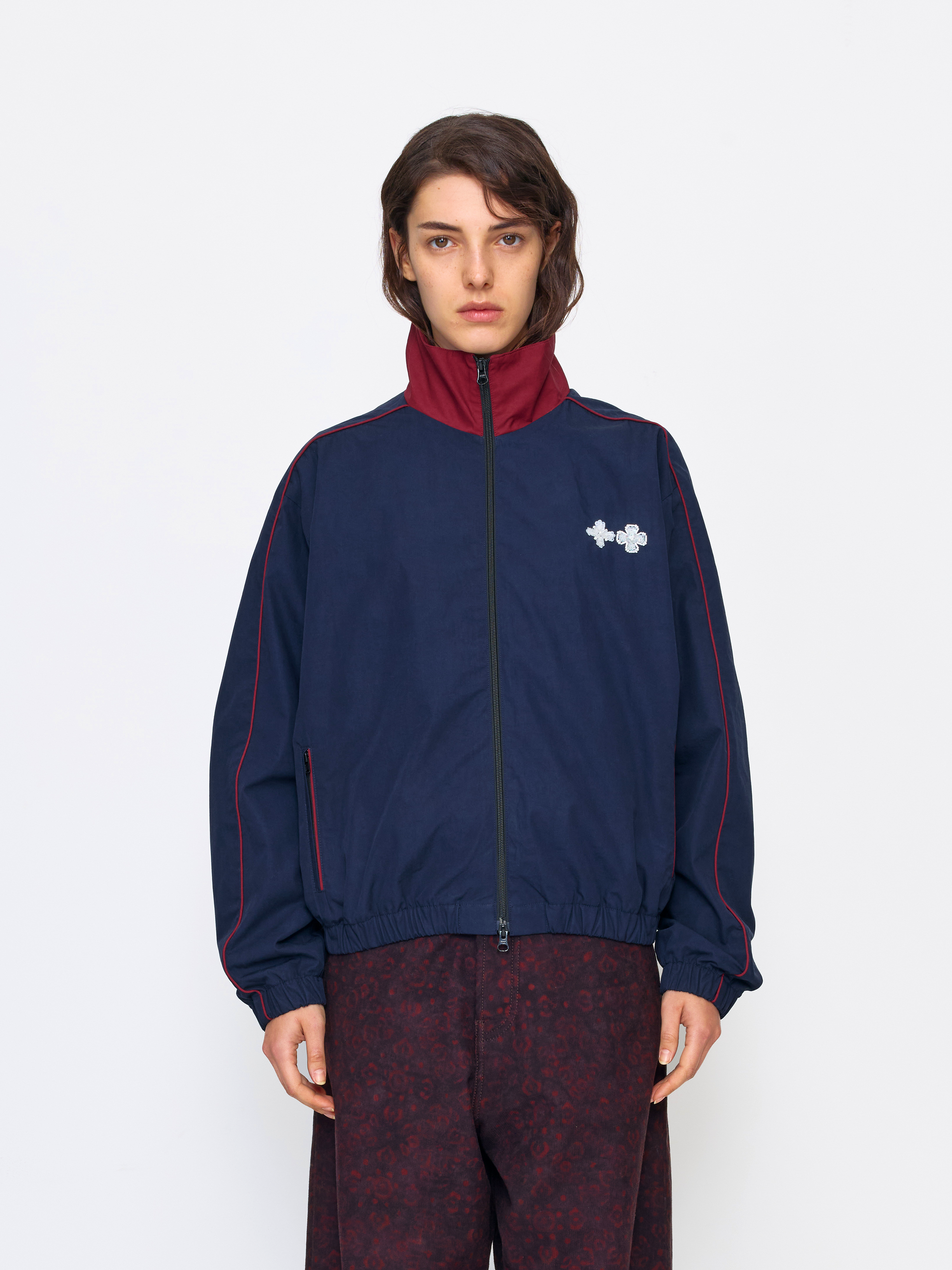 A model wearing a navy blue zip-up track jacket with a burgundy collar and red piping details, paired with burgundy patterned pants, against a plain white studio background.